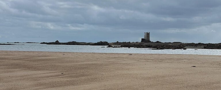Sandy beach with distant tower under cloudy sky.